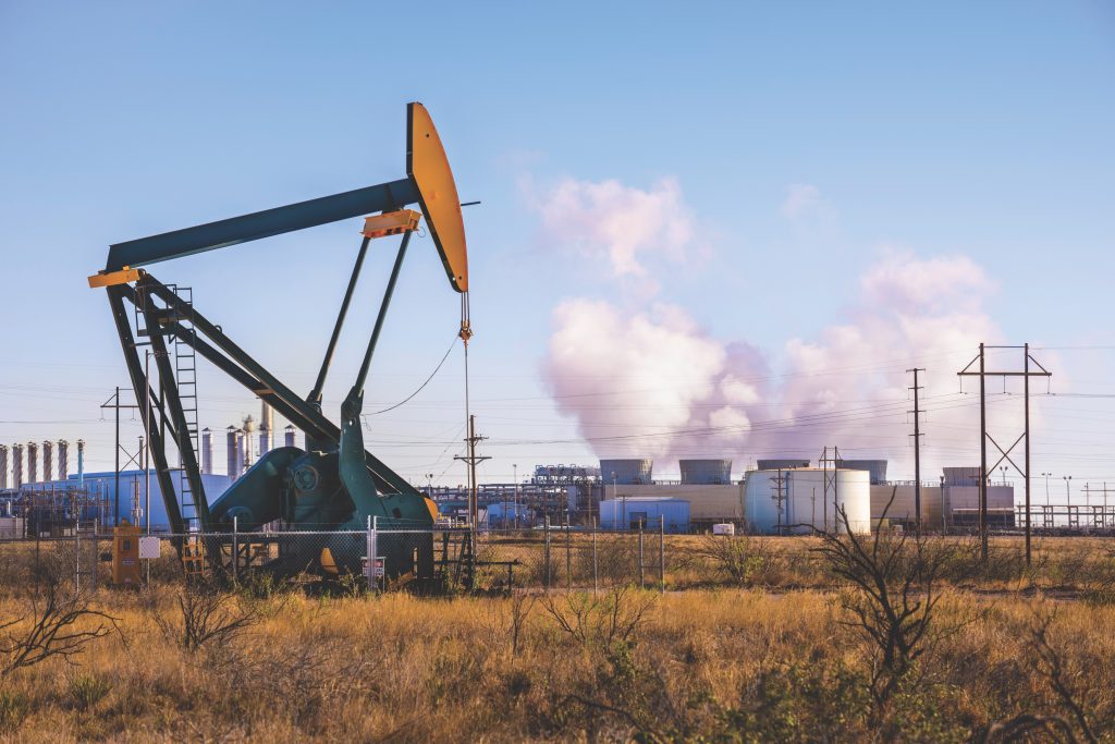 Oil pumpjack operating near an industrial refinery at sunset.