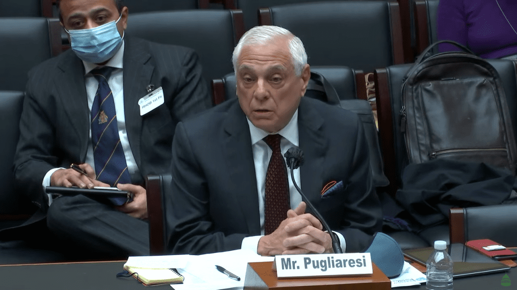 An elderly man in a suit speaking at a formal meeting.