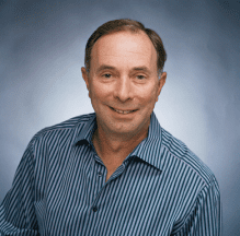 Smiling man in a blue striped shirt against a soft blue background.