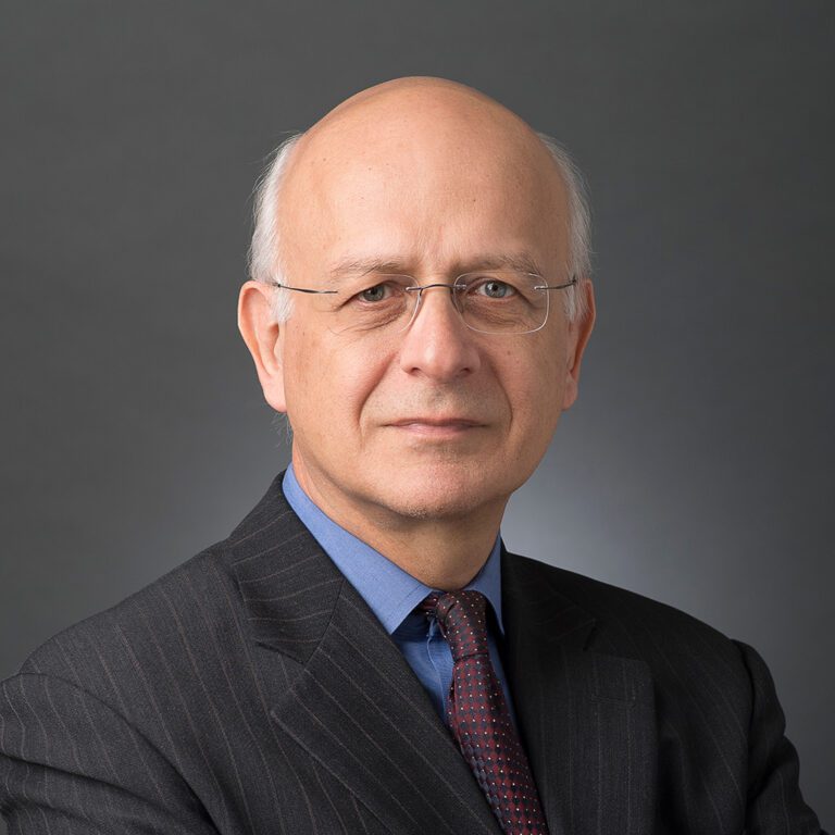 Portrait of an older man in a suit with glasses against a gray background.