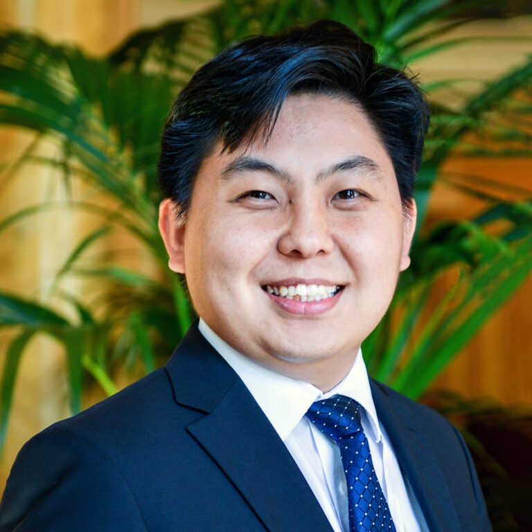 Smiling man in a suit and tie standing indoors with plants in the background.